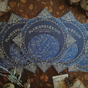 May include: A collection of dark blue Planisphaerium celestial charts with gold accents. Each chart features a detailed star map and the title "Planisphaerium" in elegant lettering. The charts are arranged on a wooden surface, evoking a vintage aesthetic.
