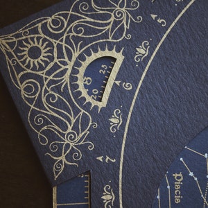 May include: Close-up of a dark blue paper with gold foil celestial design. The design includes a sun, floral patterns, and a protractor-like scale. The word "Pisces" is visible. The paper has a textured surface.