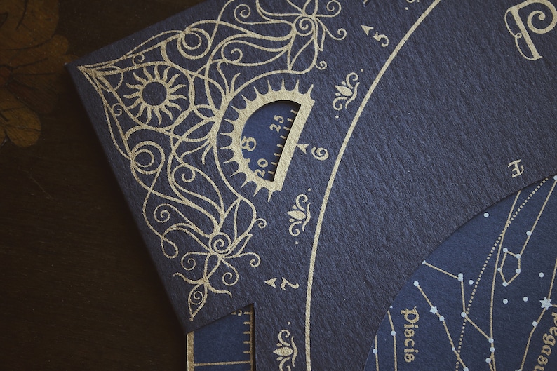 May include: Close-up of a dark blue paper with gold foil celestial design. The design includes a sun, floral patterns, and a protractor-like scale. The word "Pisces" is visible. The paper has a textured surface.