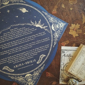 May include: A dark blue celestial map with gold accents, featuring illustrations of the sun, moon, stars, and planets. The map has decorative borders and text in multiple languages. A small book and a decorative metal pendant are also visible.