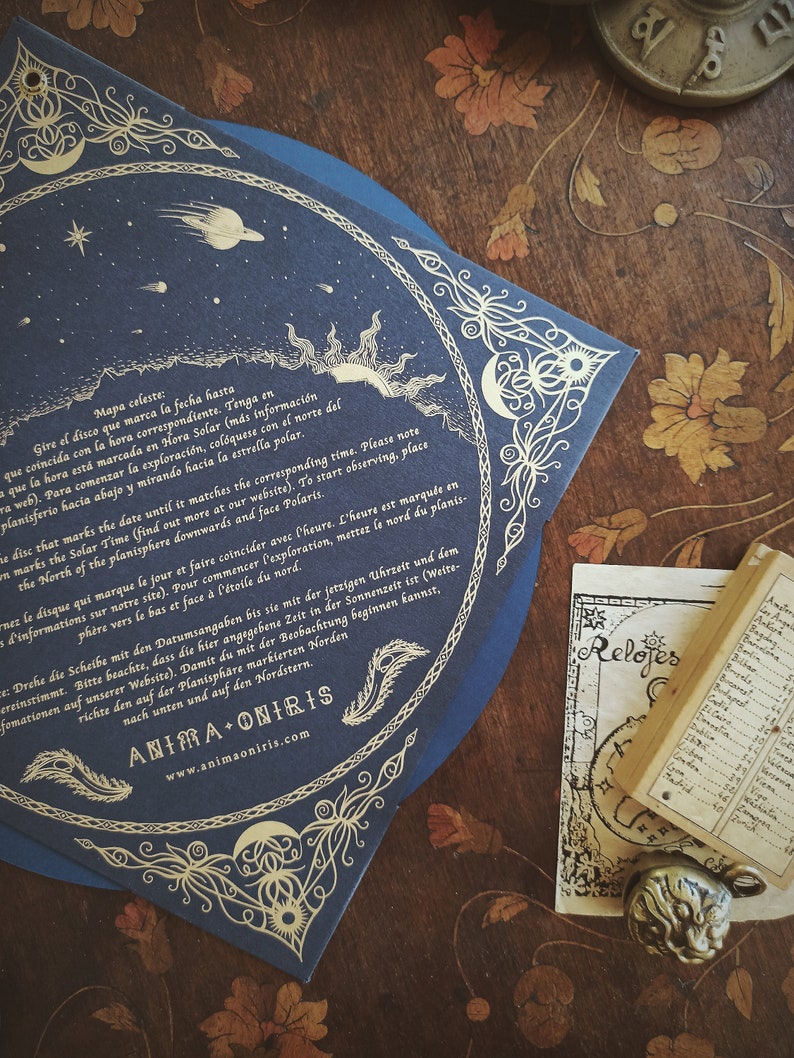 May include: A dark blue celestial map with gold accents, featuring illustrations of the sun, moon, stars, and planets. The map has decorative borders and text in multiple languages. A small book and a decorative metal pendant are also visible.