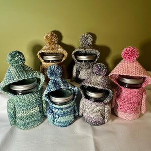 May include: A collection of six glass jars, each encased in a hand-crocheted hoodie cover featuring a hood, sleeves, and a pom-pom on top. The covers are crafted in various variegated yarn colors including green, blue, pink, grey, and gold tones.