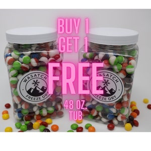 May include: Two clear plastic tubs filled with colorful freeze-dried candy. The tubs are labeled "Wasatch Freeze Dry" and the text "Buy 1 Get 1 Free 48 oz. Tub" is displayed in pink.