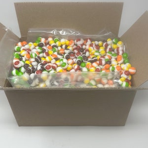 May include: A cardboard box with the lid open, revealing a clear plastic bag filled with colorful candy-coated chocolate candies. The candies are a mix of red, orange, yellow, green, brown, and white.