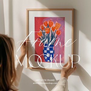 Puede incluir: Una pintura enmarcada de un ramo de tulipanes naranjas en un jarrón de cuadros azules y blancos sobre un fondo rosa. El marco es de madera natural.