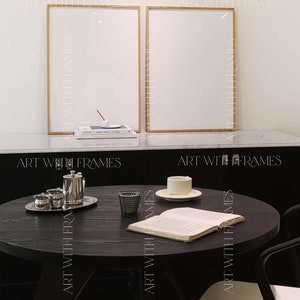 May include: A black round table with a book, a cup and saucer, and a glass on it. Two black chairs with white seats are next to the table. Two framed pictures are on the wall behind the table.