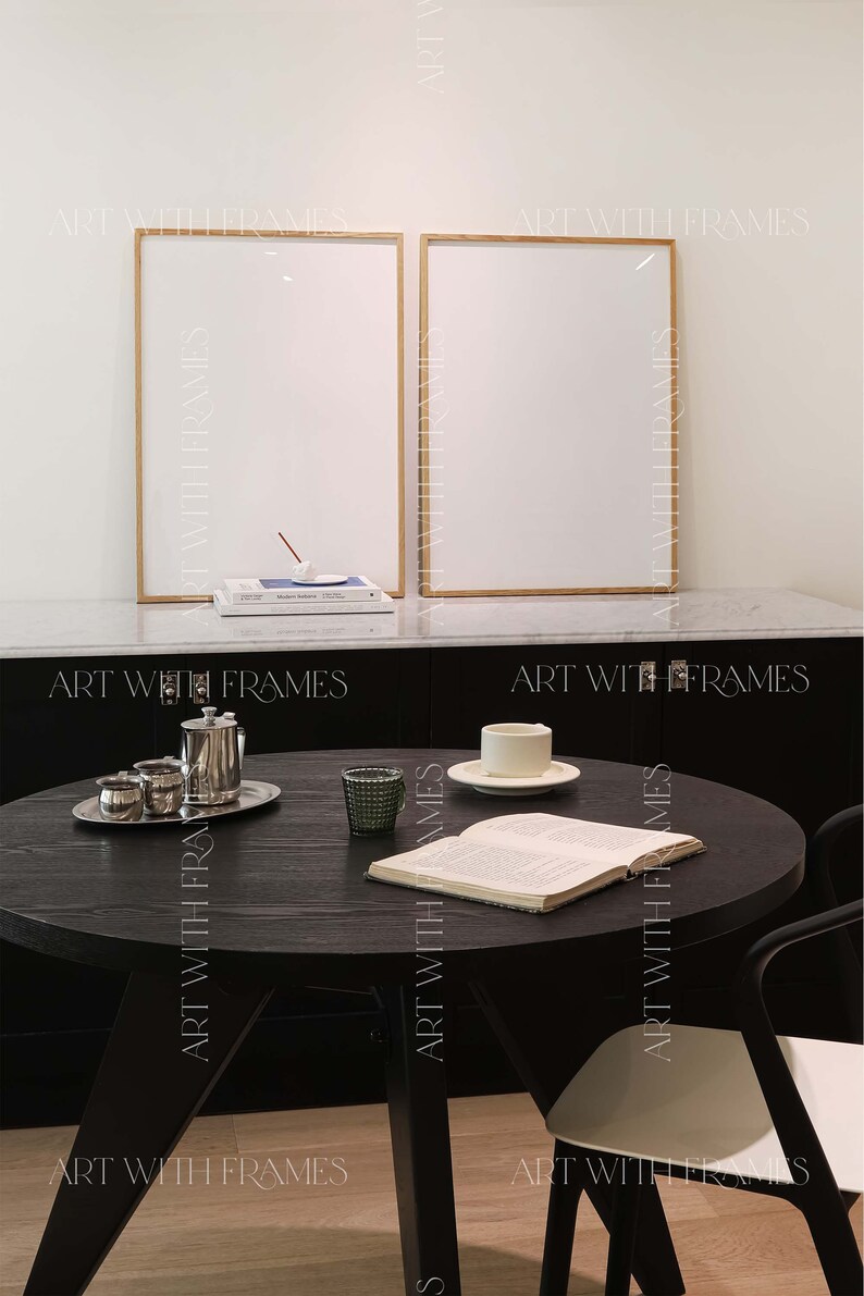 May include: A black round table with a book, a cup and saucer, and a glass on it. Two black chairs with white seats are next to the table. Two framed pictures are on the wall behind the table.