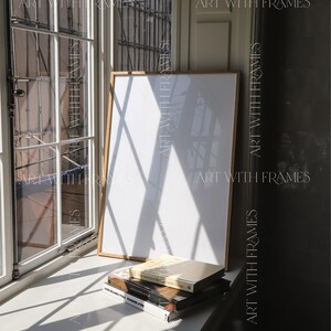 May include: A white framed picture with a white background sits on a windowsill with three books stacked on top of it. The books are titled "The Monocle Book of Gentle Living", "Scandinavian Style", and "The House That Jules Built". The windowsill is white and the background is a white wall.