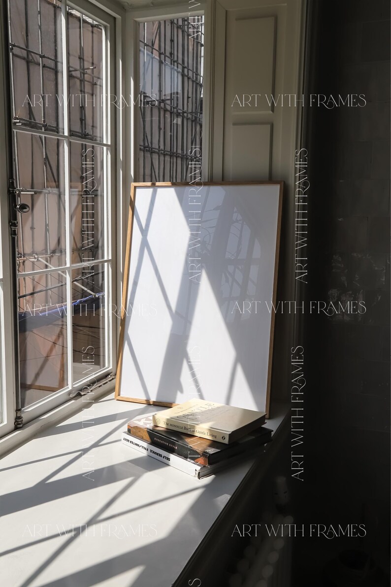 May include: A white framed picture with a white background sits on a windowsill with three books stacked on top of it. The books are titled "The Monocle Book of Gentle Living", "Scandinavian Style", and "The House That Jules Built". The windowsill is white and the background is a white wall.