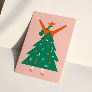 May include: A pink card with a green Christmas tree illustration. A person in a red sweater and a star on their head is standing inside the tree. The tree is decorated with ornaments, bows, and a star on top.
