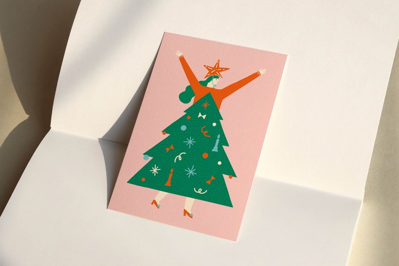 May include: A pink card with a green Christmas tree illustration. A person in a red sweater and a star on their head is standing inside the tree. The tree is decorated with ornaments, bows, and a star on top.