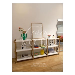 May include: A staged interior shot featuring white shelving units with wooden accents. The shelves display decorative items like a green cactus-shaped vase, a small radio, books, and a yellow decorative object. A large, blank picture frame leans against the wall.