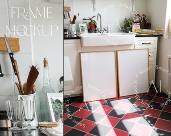 May include: Two wooden framed pictures with white mats are leaning against a white wall in a kitchen. The kitchen has a white tiled backsplash, a white sink, and a white countertop. The floor is black and red checkered tiles.