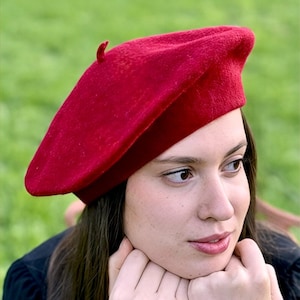 May include: A woman wearing a red beret and a black sweater, with her hands resting on her chin, looking off to the side.