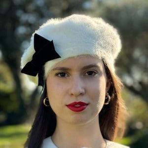 May include: A woman wearing a white fuzzy beret with a black velvet bow, a white collared shirt with a black bow and a white flower, and a black and white striped shirt.