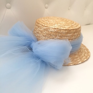 Puede incluir: Un sombrero de paja con un ala ancha y una corona plana, adornado con un lazo de tul azul claro.