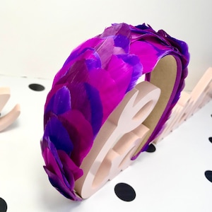 May include: A purple and pink feather headband. The headband is made of feathers that are arranged in a circular pattern. The headband is on a pink surface.