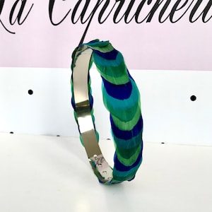 Puede incluir: Una diadema plateada con un diseño de plumas de pavo real. Las plumas son azules y verdes.