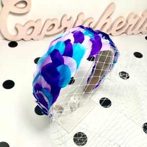 May include: A decorative headband featuring a vibrant arrangement of feathers in shades of purple, blue, and white. The feathers are attached to a headband and accented with a white netting. The word "Capricorn" is visible in the background.
