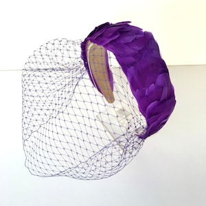May include: Purple feather headband with a matching purple birdcage veil. The headband is made of a soft, flexible material and is adorned with a cluster of feathers. The veil is made of a fine mesh material and is attached to the headband with a small comb.