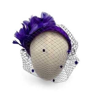 May include: A purple fascinator with a headband base, adorned with feathers and a black netting veil. The veil is dotted with small purple accents. The headpiece is displayed on a mannequin head.