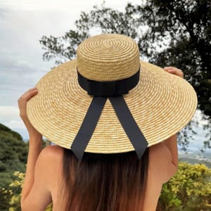 Könnte beinhalten: Ein breitkrempiger Strohhut mit runder Krone und schwarzem Schleifenband auf der Rückseite. Der Hut hat eine natürliche beige Farbe und bietet Sonnenschutz. Der Hut hat eine breite Krempe.