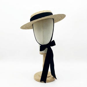 Puede incluir: Un sombrero de paja tipo canotier con una cinta de grogrén negra y una larga cinta de terciopelo negro. El sombrero se exhibe sobre una cabeza de maniquí y un soporte de madera sobre un fondo blanco. Un sombrero clásico.