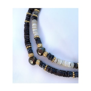 Puede incluir: Un collar de capas con cuentas de concha marrón y blanca, detalles dorados y una piedra preciosa gris oscura.