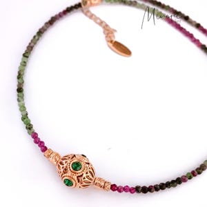 May include: A delicate necklace featuring a central filigree pendant with green gemstones, flanked by strands of small, colorful beads. The beads transition from green to pink and dark hues. The necklace has a gold-toned clasp and a small tag.