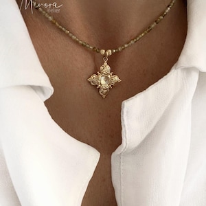 May include: A gold necklace with a delicate chain and a small, ornate cross pendant. The necklace is made of tiny brown beads and is worn by a person with a white shirt.