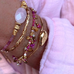 Peut inclure: Plusieurs bracelets dorés, dont un torsadé et d'autres ornés de perles roses et dorées. L'un des bracelets présente une grosse pierre précieuse ovale rose pâle. Un charm en forme de coquillage doré.
