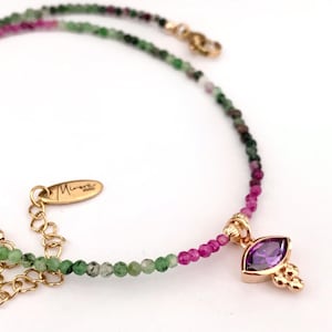 Op de afbeelding: Een delicate gouden ketting met een gradiënt van groene en roze kralen. Een klein hanger met een paarse edelsteen hangt aan de ketting.