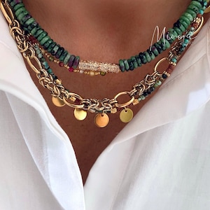 Op de afbeelding: Een gelaagde ketting set. De bovenste laag is een groene kralenketting, gevolgd door een gouden ketting met kleine schijfjes en een gouden ketting met een decoratieve staaf. De kettingen worden getoond tegen een wit overhemd.
