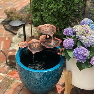 Puede incluir: Fuente de agua decorativa para exteriores con detalles de nenúfares de color cobre. La fuente está en una maceta de cerámica turquesa con un diseño floral. Una maceta de hortensias moradas y azules está al lado.