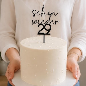 Może przedstawiać: Czarny topper na tort z niemieckim napisem "schon wieder 29" wykonanym kursywą i pogrubioną czcionką, umieszczony na cylindrycznym białym torcie ozdobionym małymi białymi jadalnymi perełkami. Topper posiada cienki szpikulec do wbicia w ciasto.