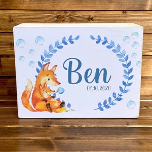 Pode incluir: Uma placa de madeira branca com uma grinalda floral azul e branca, duas raposas e o nome "Ben" com a data "01.10.2020".