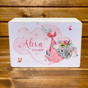 Puede incluir: Una caja de madera blanca con una ilustración rosa y gris de un elefante que lleva una cigüeña. La caja tiene el nombre "Alexa" y la fecha "03.03.2007" escrita en ella.