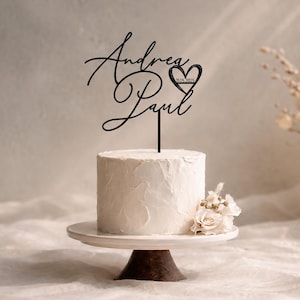 Op de afbeelding: Een zwarte taarttopper met de namen Andrea en Paul in een cursief lettertype, inclusief een hartvorm met de datum 19.05.2025. De topper staat bovenop een kleine, witte taart met een textuur, geplaatst op een witte taartstandaard.
