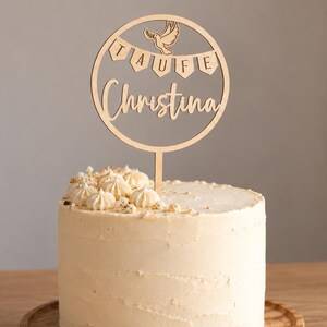Può includere: Un topper per torta rotondo in legno con una colomba tagliata al laser, uno stendardo con il testo TAUFE e il nome Christina in corsivo, posizionato sopra una torta cilindrica con glassa chiara e piccole decorazioni sulla parte superiore.