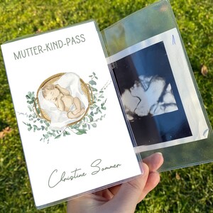 May include: A clear plastic document holder with a printed cover featuring the text "MUTTER-KIND-PASS" and a watercolor illustration of a baby in a basket. The holder contains an ultrasound image.