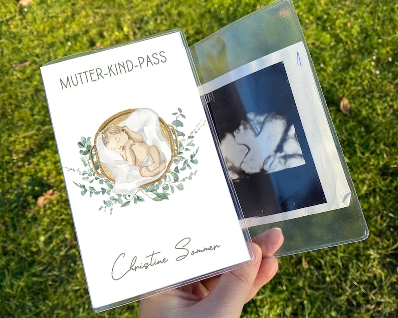 May include: A clear plastic document holder with a printed cover featuring the text "MUTTER-KIND-PASS" and a watercolor illustration of a baby in a basket. The holder contains an ultrasound image.
