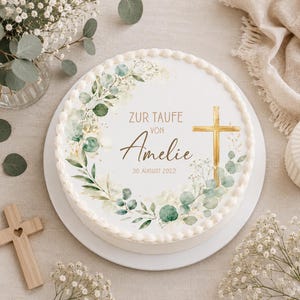 Peut inclure: Un disque décoratif pour gâteau orné d'une couronne d'eucalyptus à l'aquarelle, d'une croix dorée et du texte ZUR TAUFE VON Amelie 30 AUGUST 2022. Le motif présente des feuilles vertes et des fleurs blanches sur un fond blanc bordé de crème.
