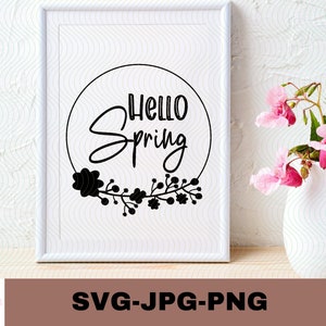 May include: A white framed print with a black circle containing the text "Hello Spring" and a floral wreath. The print is on a white wall with a vase of pink flowers in the foreground.