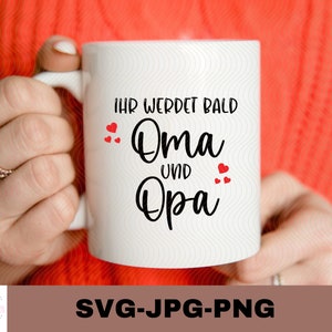 May include: White ceramic mug with black text that reads "Ihr werdet bald Oma und Opa" with red hearts around the text.