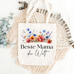 May include: A white canvas tote bag with a floral design and the text "Beste Mama der Welt" in black. The bag has a long handle and is made of natural fabric.