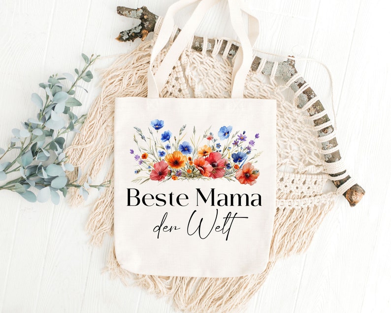 May include: A white canvas tote bag with a floral design and the text "Beste Mama der Welt" in black. The bag has a long handle and is made of natural fabric.