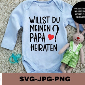 May include: A light blue baby onesie with the text "WILLST DU MEINEN PAPA? HEIRATEN" in black with a red heart. The onesie is on a white fuzzy surface with a stuffed bunny toy.