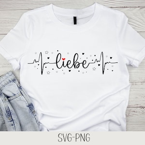 Peut inclure: T-shirt blanc avec un graphique de battement de cœur noir et rouge qui dit "liebe" en écriture noire. Le graphique est entouré de petites étoiles et de cœurs noirs.