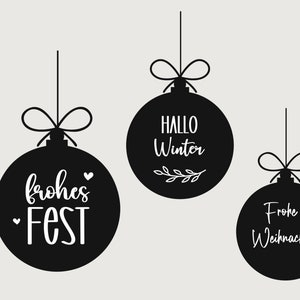 Peut inclure: Trois boules de Noël noires avec du texte blanc. La première boule dit "Hallo Winter" avec un petit motif de branche. La deuxième boule dit "Frohes Fest" avec un petit cœur. La troisième boule dit "Frohe Weihnachten".