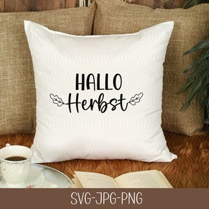 May include: White cushion with black text that says "Hallo Herbst" with two leaf designs on either side of the text. The cushion is on a brown fluffy surface with a book and a cup of coffee.